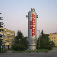 Wysoki, srebrny zbiornik z napisem RAFAKO w czerwonych literach, otoczony zielenią i budynkami fabrycznymi.