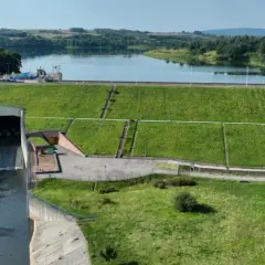 Zapora wodna z budynkiem elektrowni i kanałem odpływowym na tle jeziora i lasów.
