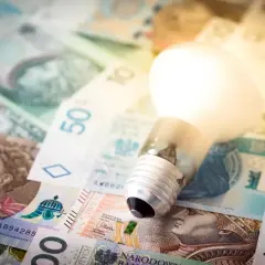 Żarówka na stosie polskich banknotów symbolizuje koszty energii, np. taryfa G11 Tauron.