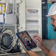Technik w kasku i okularach ochronnych wykonuje przegląd instalacji elektrycznej, korzystając z tabletu z aplikacją diagnostyczną.