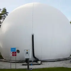 Ogromny, biały balon magazynujący biometan, otoczony drzewami. Obok znajduje się panel sterowania i rury.
