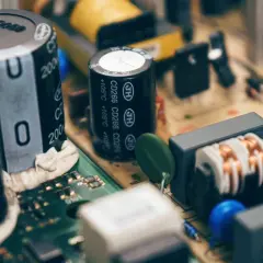 Zbliżenie na budowę kondensatora i innych elementów elektronicznych na płytce drukowanej. Widać cewki, rezystory i niebieskie oraz żółte elementy.