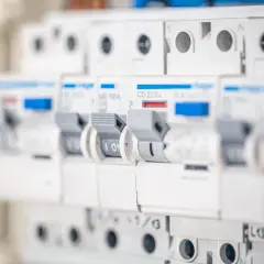Szukasz informacji, jaki bezpiecznik do gniazdek? Na zdjęciu widać rząd białych wyłączników nadprądowych w rozdzielnicy elektrycznej, każdy z dźwignią i oznaczeniem prądowym.