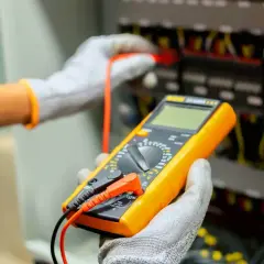 Elektryk w rękawicach dokonuje odbioru instalacji elektrycznej, używając multimetru do sprawdzenia parametrów.
