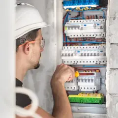 Elektryk w kasku i okularach ochronnych pracuje przy skrzynce z bezpiecznikami.