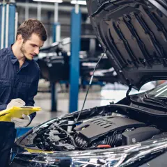 Mechanik w granatowym kombinezonie notuje coś na żółtej teczce, stojąc przy otwartej masce samochodu. To skrót do świata koni mechanicznych.