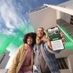 Dwie uśmiechnięte kobiety trzymają telefon z ofertą pracy w Fortum. Zielone fale energii otaczają budynki, symbolizując moc i rozwój.