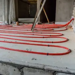 Układanie rur ogrzewania podłogowego na styropianie. Widać czerwone rurki ułożone w pętle na betonowej płycie, gotowe do zalania.
