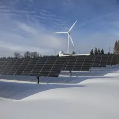 Panele słoneczne i wiatrak na ośnieżonym polu. W tle las i budynek. To obraz przyszłości, gdzie opłata sieciowa zmienna całodobowa wspiera zieloną energię.