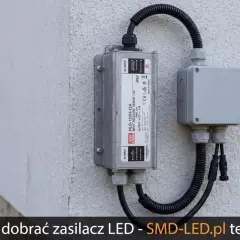 Zasilacz LED zamontowany na ścianie z podłączonymi przewodami w peszlu do puszki instalacyjnej.