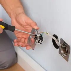 Ręce majsterkowicza przykręcają śrubokrętem mechanizm gniazdka. Widać przewody elektryczne wychodzące ze ściany.