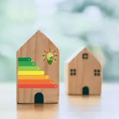 Drewniane domki z etykietą energetyczną, gdzie jeden z nich ma klasę energetyczną G. Symbol żarówki z liściem sugeruje ekologiczne rozwiązania.