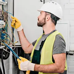 Elektryk w kasku i kamizelce odblaskowej dokonuje wymiany instalacji elektrycznej, pracując przy skrzynce z przewodami.