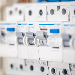 Szukasz informacji, jaki bezpiecznik do gniazdek? Na zdjęciu widać rząd białych wyłączników nadprądowych w rozdzielnicy elektrycznej, każdy z dźwignią i oznaczeniem prądowym.
