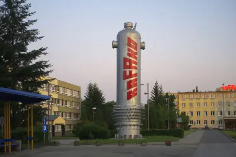 Wysoki, srebrny zbiornik z napisem RAFAKO w czerwonych literach, otoczony zielenią i budynkami fabrycznymi.
