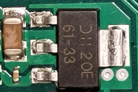 Czarny element z oznaczeniem "DH 20E 61-33" to elektroda tranzystora, otoczona innymi komponentami na zielonej płytce drukowanej.