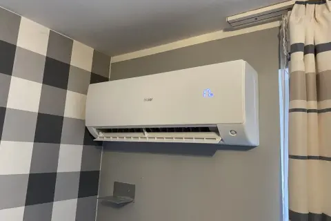 Biała jednostka klimatyzacyjna Haier zamontowana na szarej ścianie, obok tapety w kratę i zasłon.