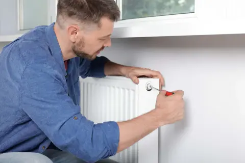 Mężczyzna używa śrubokręta, by odpowietrzyć grzejnik. To prosty sposób na pozbycie się powietrza z instalacji grzewczej.