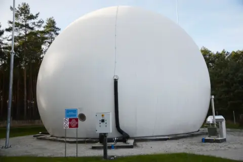Ogromny, biały balon magazynujący biometan, otoczony drzewami. Obok znajduje się panel sterowania i rury.