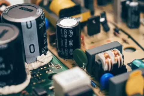 Zbliżenie na budowę kondensatora i innych elementów elektronicznych na płytce drukowanej. Widać cewki, rezystory i niebieskie oraz żółte elementy.