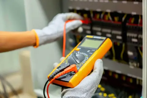 Elektryk w rękawicach dokonuje odbioru instalacji elektrycznej, używając multimetru do sprawdzenia parametrów.