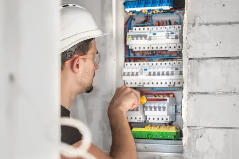Elektryk w kasku i okularach ochronnych pracuje przy skrzynce z bezpiecznikami.