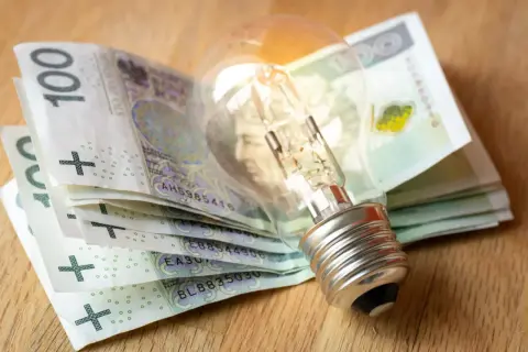 Żarówka na stosie banknotów 100 zł symbolizuje wysokie koszty energii. Dodatek energetyczny może pomóc w pokryciu rachunków.