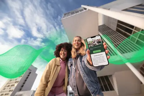Dwie uśmiechnięte kobiety trzymają telefon z ofertą pracy w Fortum. Zielone fale energii otaczają budynki, symbolizując moc i rozwój.