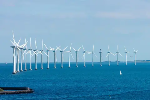 Rząd białych turbin wiatrowych tworzy imponującą farmę wiatrową na Bałtyku. Na horyzoncie widać żaglówkę i samolot.