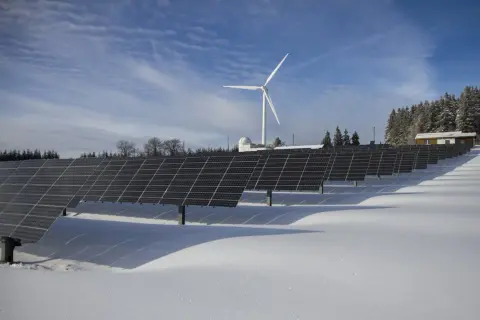 Panele słoneczne i wiatrak na ośnieżonym polu. W tle las i budynek. To obraz przyszłości, gdzie opłata sieciowa zmienna całodobowa wspiera zieloną energię.