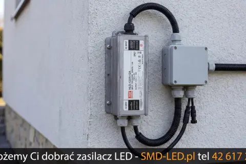 Zasilacz LED zamontowany na ścianie z podłączonymi przewodami w peszlu do puszki instalacyjnej.
