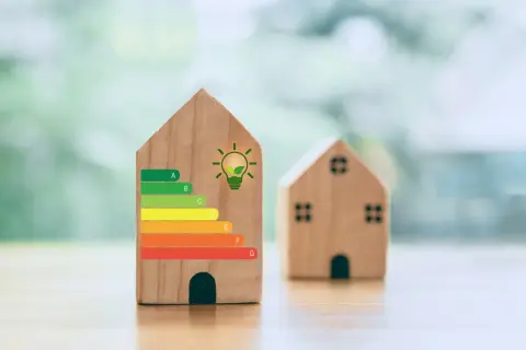 Drewniane domki z etykietą energetyczną, gdzie jeden z nich ma klasę energetyczną G. Symbol żarówki z liściem sugeruje ekologiczne rozwiązania.