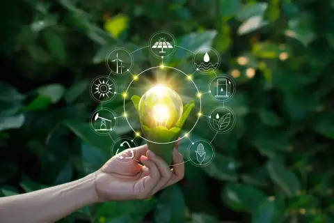 Dłoń trzyma żarówkę z zielonymi liśćmi, otoczoną ikonami symbolizującymi różne źródła energii, w tym OZE.