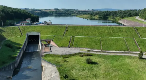 Zapora wodna z budynkiem elektrowni i kanałem odpływowym na tle jeziora i lasów.