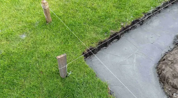 Wyznaczanie fundamentów domu. Drewniane paliki i biała nić wyznaczają linię wykopu pod fundamenty, obok świeżo wylanej ławy betonowej.