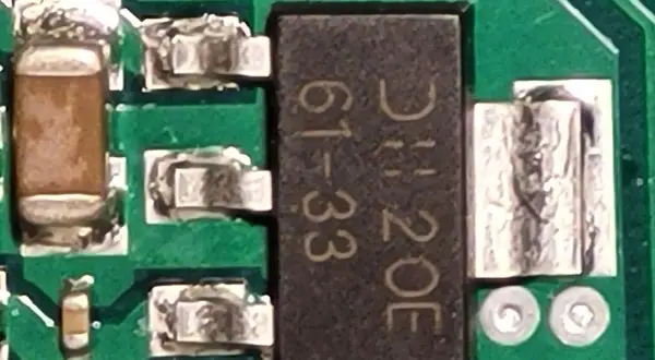 Czarny element z oznaczeniem "DH 20E 61-33" to elektroda tranzystora, otoczona innymi komponentami na zielonej płytce drukowanej.