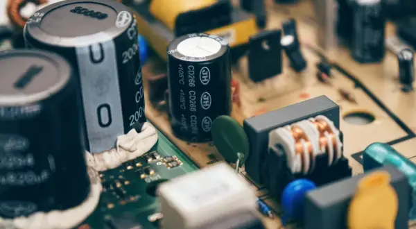 Zbliżenie na budowę kondensatora i innych elementów elektronicznych na płytce drukowanej. Widać cewki, rezystory i niebieskie oraz żółte elementy.