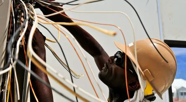 Elektryk w kasku i okularach pracuje przy instalacji elektrycznej, łącząc przewody. To dobry przykład, jak zostać elektrykiem.