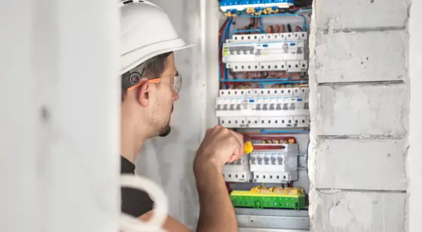 Elektryk w kasku i okularach ochronnych pracuje przy skrzynce z bezpiecznikami.