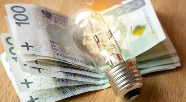 Żarówka na stosie banknotów 100 zł symbolizuje wysokie koszty energii. Dodatek energetyczny może pomóc w pokryciu rachunków.