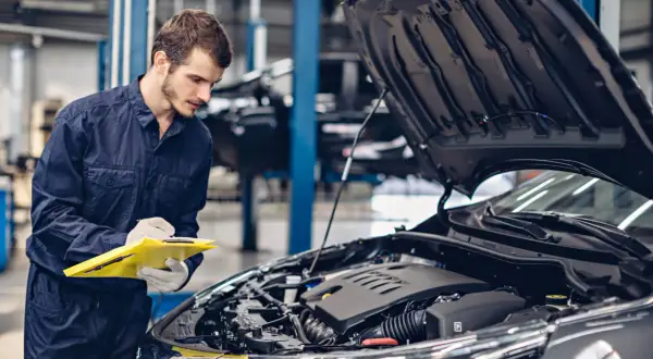 Mechanik w granatowym kombinezonie notuje coś na żółtej teczce, stojąc przy otwartej masce samochodu. To skrót do świata koni mechanicznych.