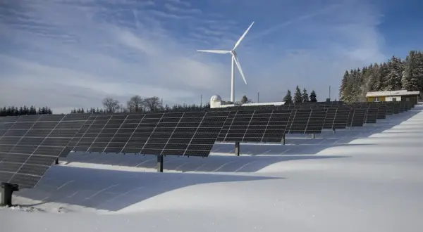 Panele słoneczne i wiatrak na ośnieżonym polu. W tle las i budynek. To obraz przyszłości, gdzie opłata sieciowa zmienna całodobowa wspiera zieloną energię.