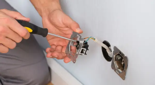Ręce majsterkowicza przykręcają śrubokrętem mechanizm gniazdka. Widać przewody elektryczne wychodzące ze ściany.
