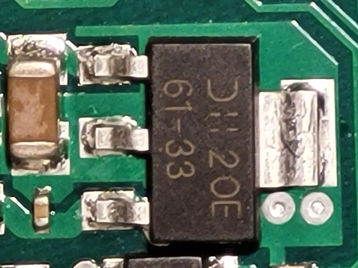 Czarny element z oznaczeniem "DH 20E 61-33" to elektroda tranzystora, otoczona innymi komponentami na zielonej płytce drukowanej.