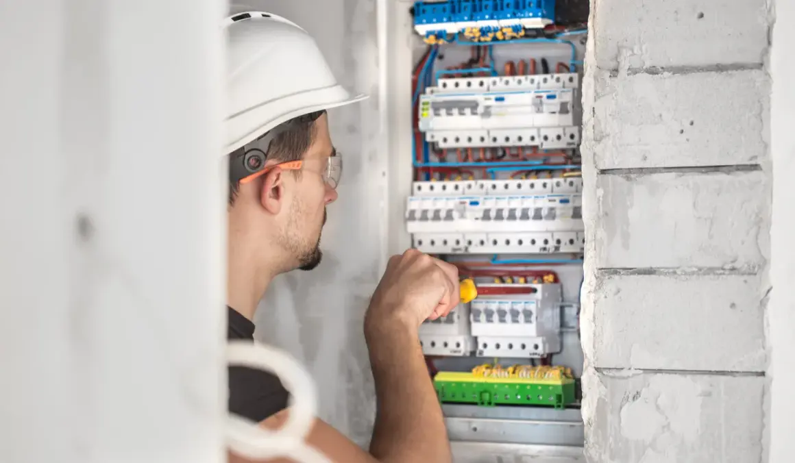 Elektryk w kasku i okularach ochronnych pracuje przy skrzynce z bezpiecznikami.