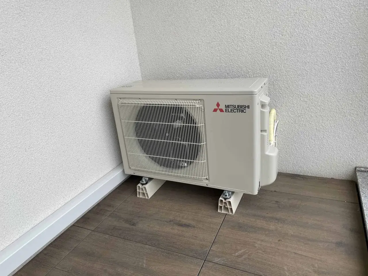Jednostka zewnętrzna klimatyzatora Mitsubishi Electric na drewnianej podłodze przy białej ścianie.