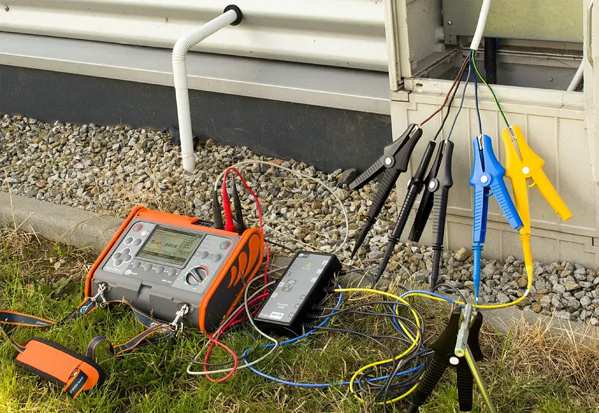 Pomiar rezystancji izolacji instalacji elektrycznej za pomocą miernika. Podłączone przewody z krokodylkami do kabli wychodzących z budynku.