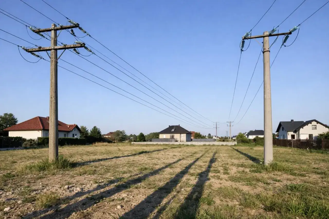Dwa słupy średniego napięcia przecinają niebo nad pustą działką. W tle widać domy mieszkalne.