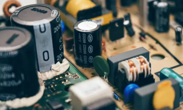 Zbliżenie na budowę kondensatora i innych elementów elektronicznych na płytce drukowanej. Widać cewki, rezystory i niebieskie oraz żółte elementy.