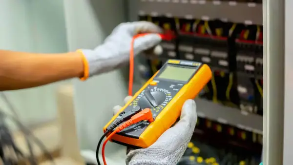 Elektryk w rękawicach dokonuje odbioru instalacji elektrycznej, używając multimetru do sprawdzenia parametrów.